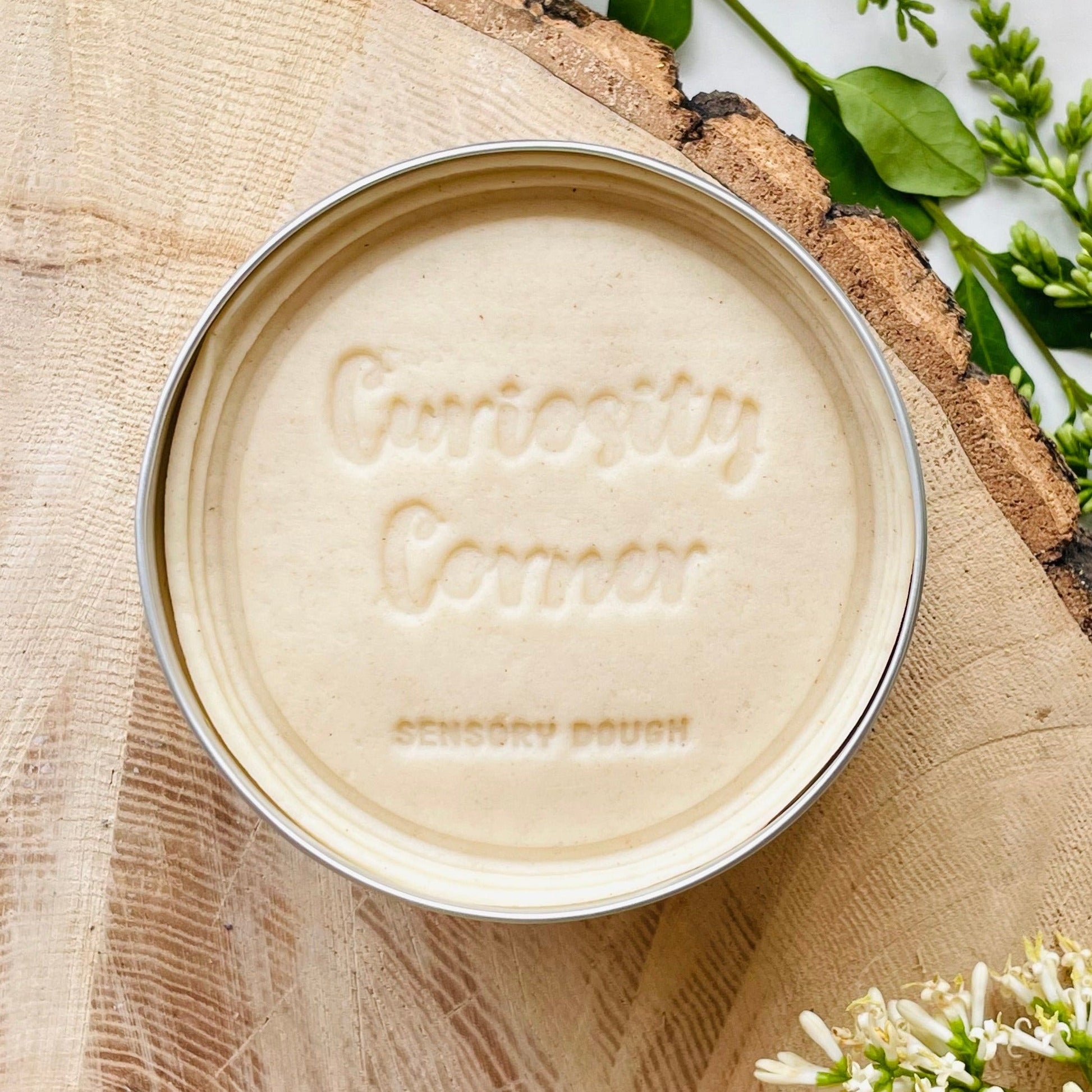 Tin of Curiosity Corner Frost White dough on a wooden surface with greenery